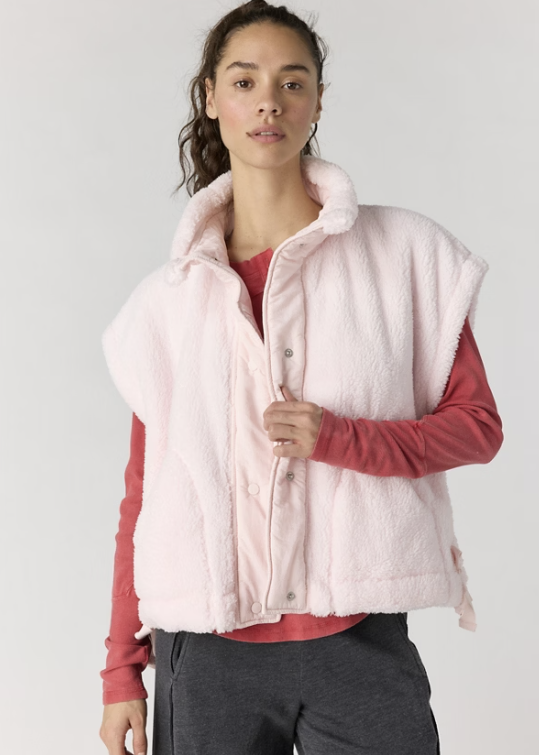 A woman wearing a light pink fuzzy sleeveless button-front vest over a dusty rose long-sleeve top and dark gray pants, photographed against a plain light gray studio background.