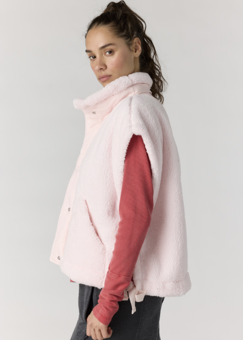 Woman wearing light pink sherpa vest over red long sleeve with gray pants on plain background