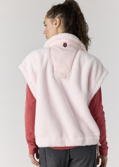 Woman shown from back wearing light pink sherpa vest with red long sleeve and gray pants on plain background