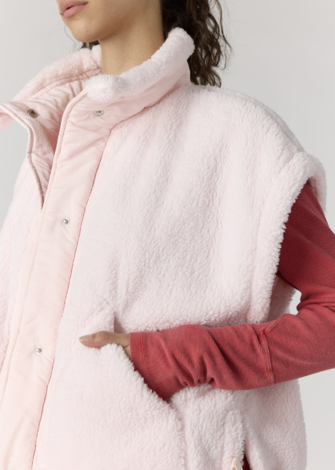 A close-up of a person wearing a pale pink sherpa fleece vest over a muted red long-sleeve shirt. The vest features a high plush collar, silver snap buttons, and side pockets, shown against a plain light gray background.