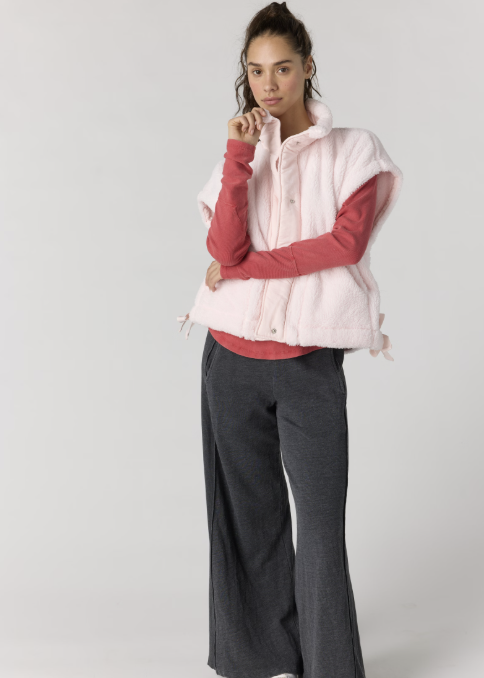 A model wearing a light pink fuzzy sleeveless vest over a coral pink long-sleeve shirt, paired with dark gray wide-leg pants and white sneakers, against a light gray studio background.