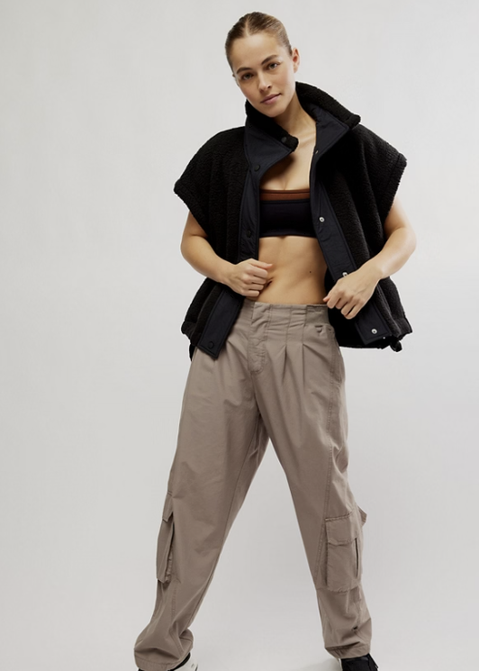 A model wearing a black fleece vest, taupe cargo pants, a black sports bra, and black sneakers, posing against a light gray studio background.