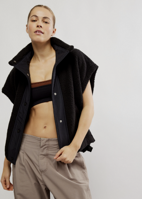 A model wearing a black sleeveless sherpa-lined zip jacket vest over a black sports bra and beige wide-leg trousers, photographed against a light gray studio background.