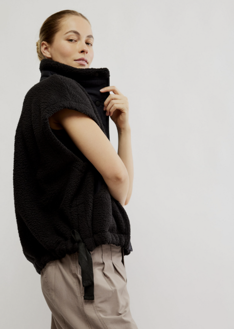 A model wearing a black sleeveless sherpa fleece vest with a high mock neck and drawstring details, paired with beige trousers, photographed against a plain light gray studio background.