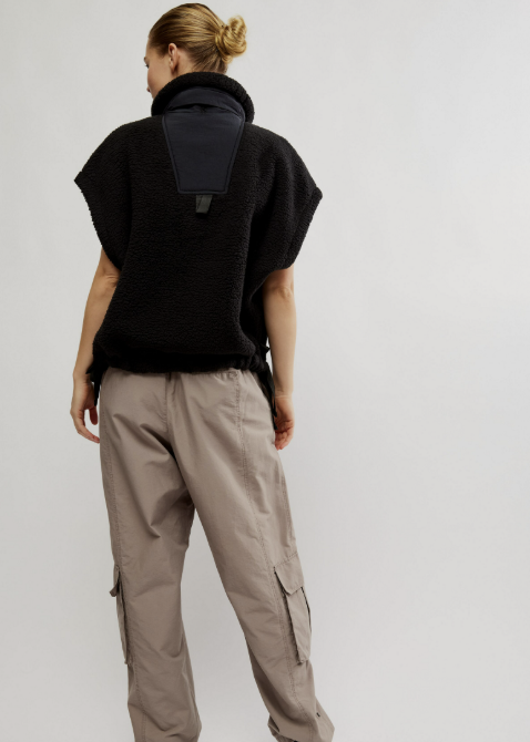 A model shown from the back wearing a black fleece pullover with a high collar and a large dark panel on the back, paired with beige cargo pants and black sneakers, against a light gray studio background.