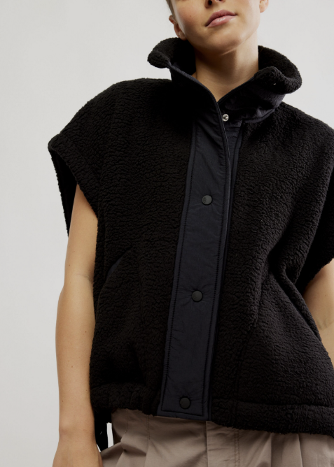 A close-up of a model wearing a black sleeveless sherpa fleece vest with a dark navy button-front placket and a high collar, paired with beige trousers, against a plain light gray background.