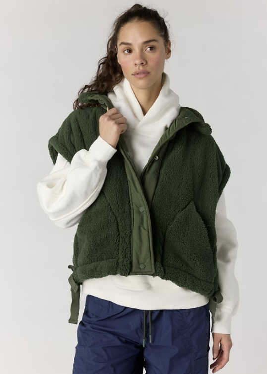 Woman wearing green fluffy vest over white hoodie and blue pants on plain background