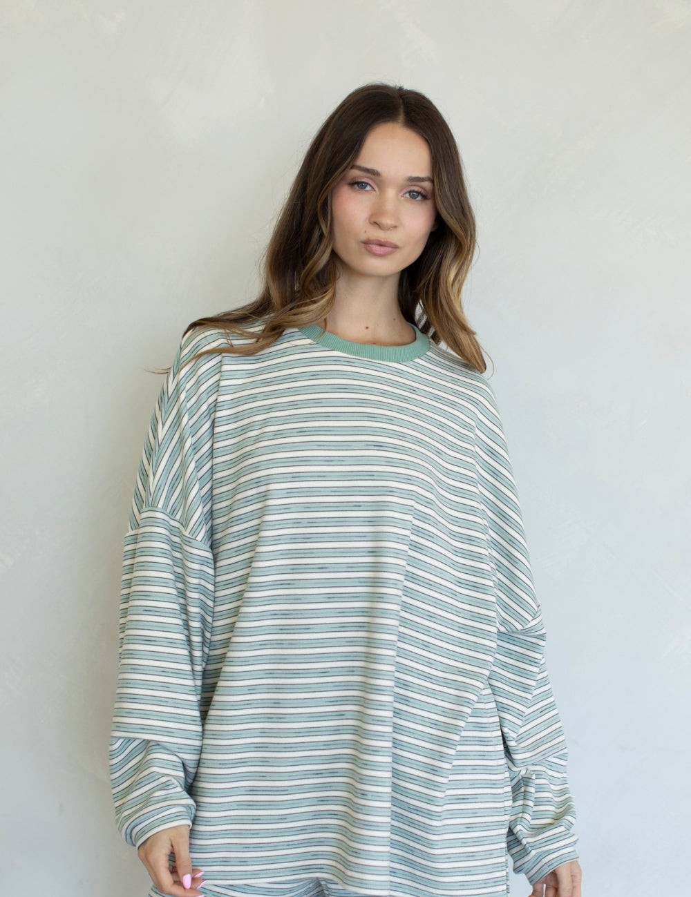 A woman modeling a loose, oversized long-sleeve top with thin horizontal stripes in sage green and white, paired with matching sage green shorts, set against a plain light gray wall.