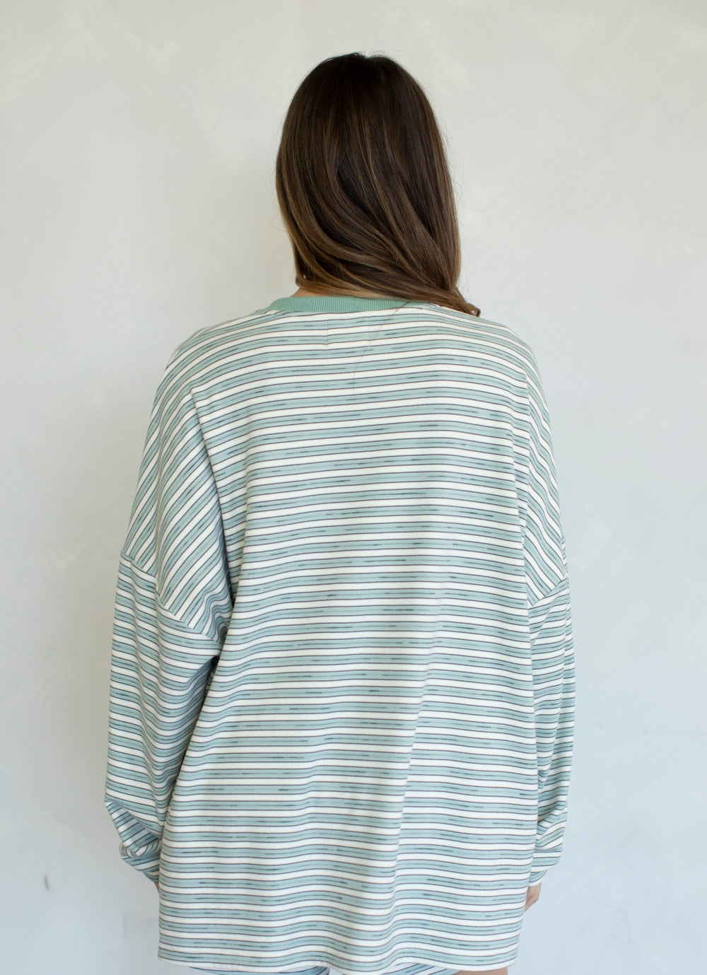 A woman viewed from behind wearing an oversized long-sleeve striped shirt with thin green and white horizontal stripes, set against a plain light gray wall.