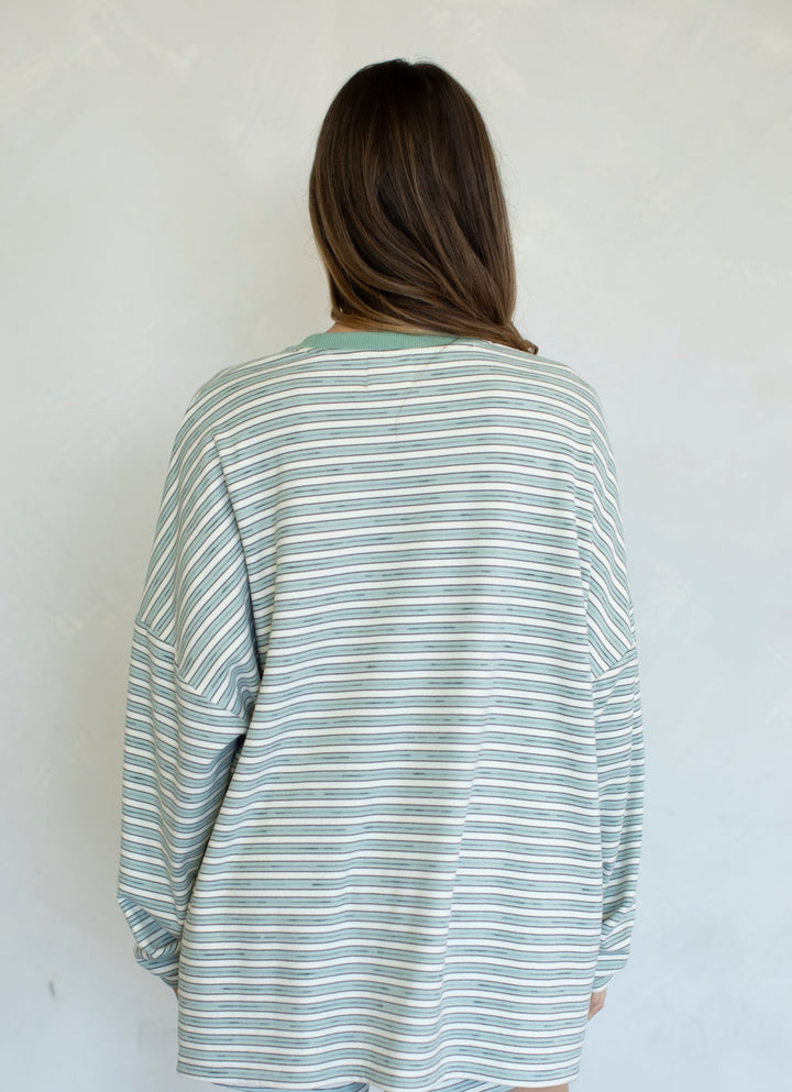 A woman viewed from behind wearing an oversized long-sleeve striped shirt with thin green and white horizontal stripes, set against a plain light gray wall.