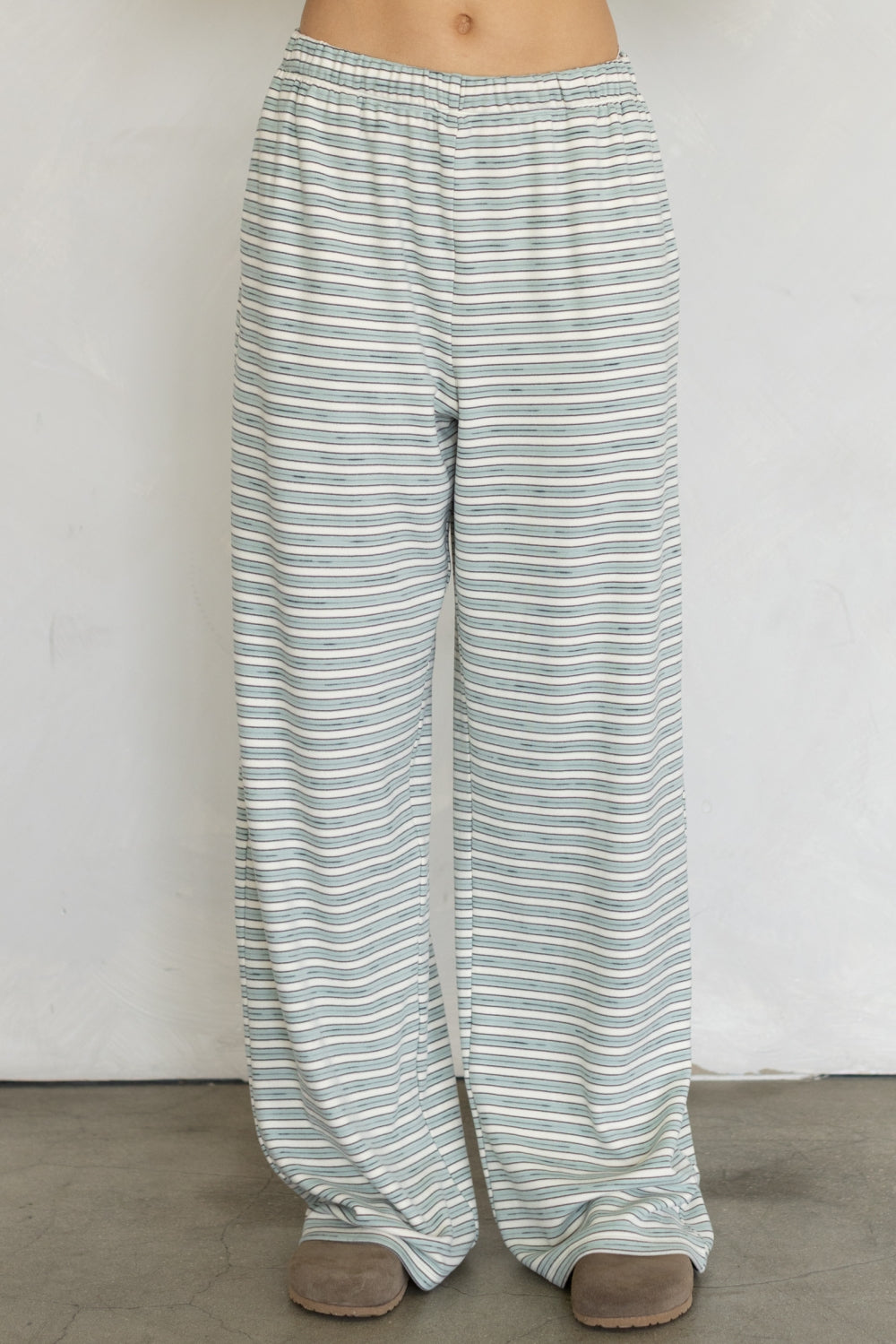 A person wearing loose, wide-leg striped lounge pants in a light sage green and white pattern, paired with brown slip-on shoes, standing against a plain off-white wall.
