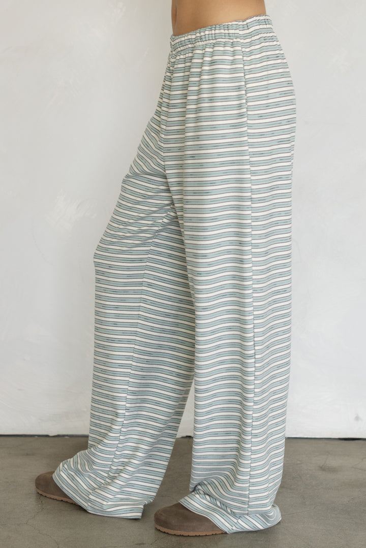 A person wearing loose, wide-leg striped lounge pants in a light sage green and white pattern, paired with brown slippers, standing against a plain off-white wall.
