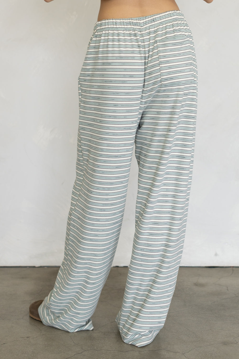A person wearing loose, wide-leg pants with thin horizontal stripes in sage green and white, shown from the waist down against a plain light wall and concrete floor.