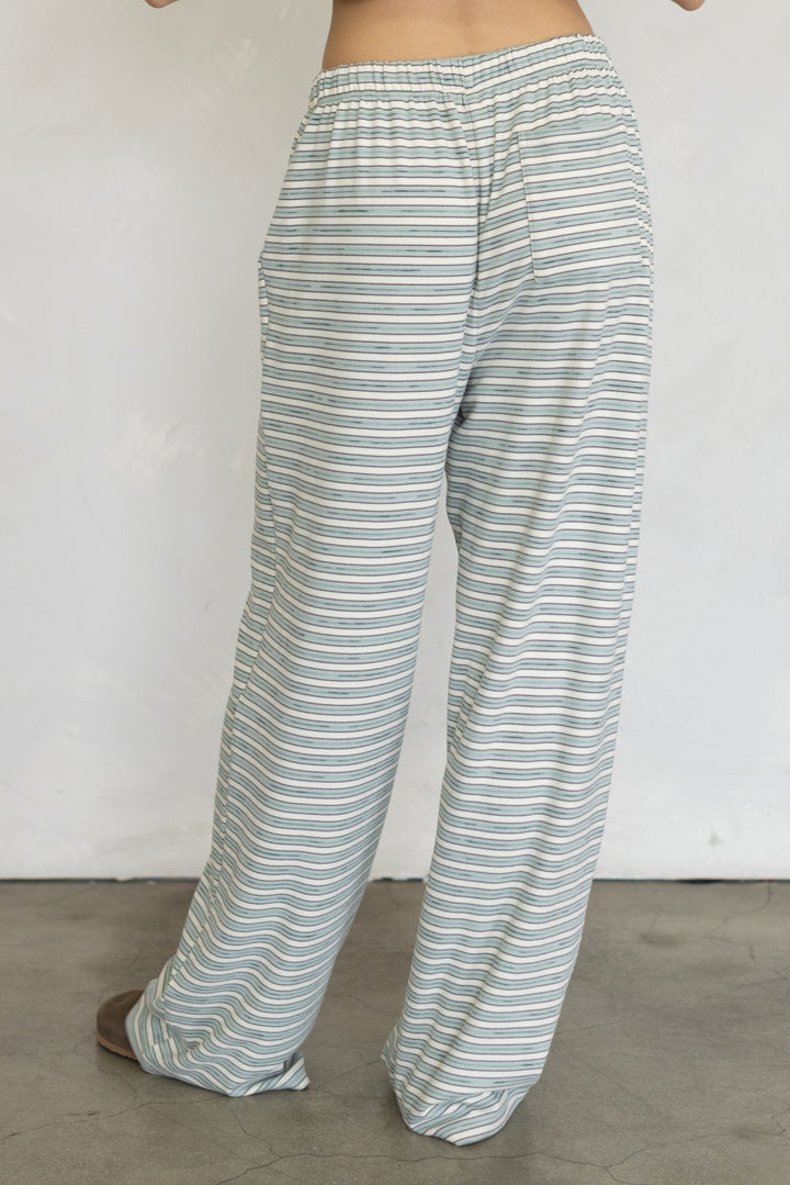 A person wearing loose, wide-leg pants with thin horizontal stripes in sage green and white, shown from the waist down against a plain light wall and concrete floor.