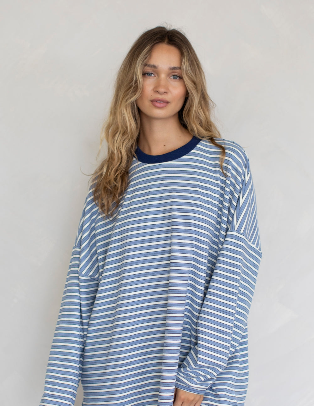 A woman modeling a long-sleeve crewneck shirt with thin blue and white horizontal stripes and a solid navy blue collar, paired with dark pants against a light gray wall.