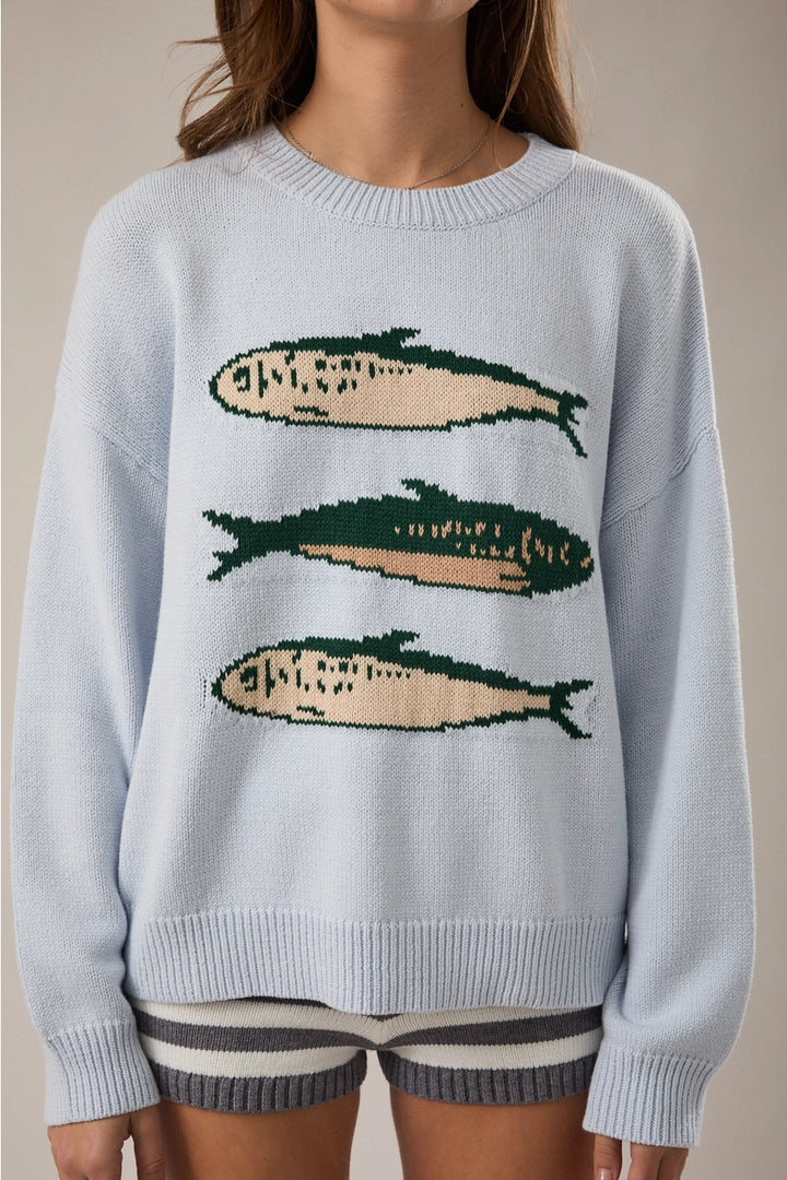 Woman wearing light blue sweater with sardine graphic on plain background