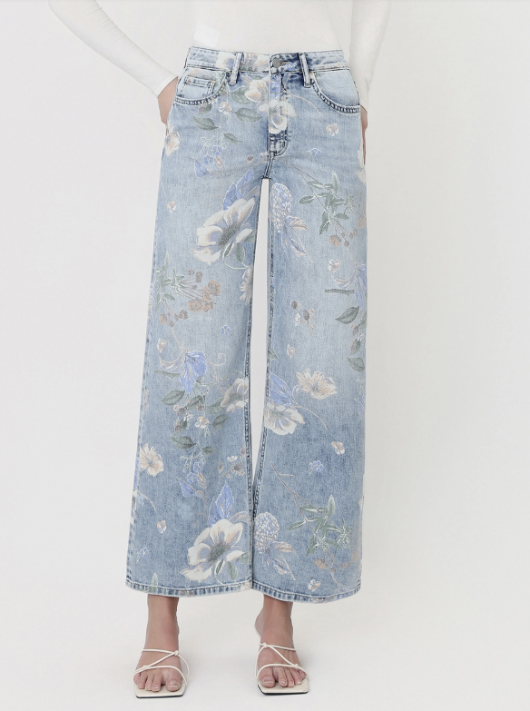 A model is wearing a pair of wide-leg light blue denim jeans with a delicate floral print, styled with a white long-sleeve top and white strappy flat sandals against a plain white studio background.