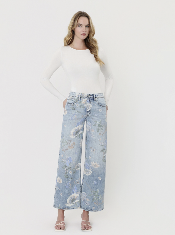 A woman stands against a plain light background wearing a fitted white long-sleeve top and light blue wide-leg floral jeans with white heeled sandals.