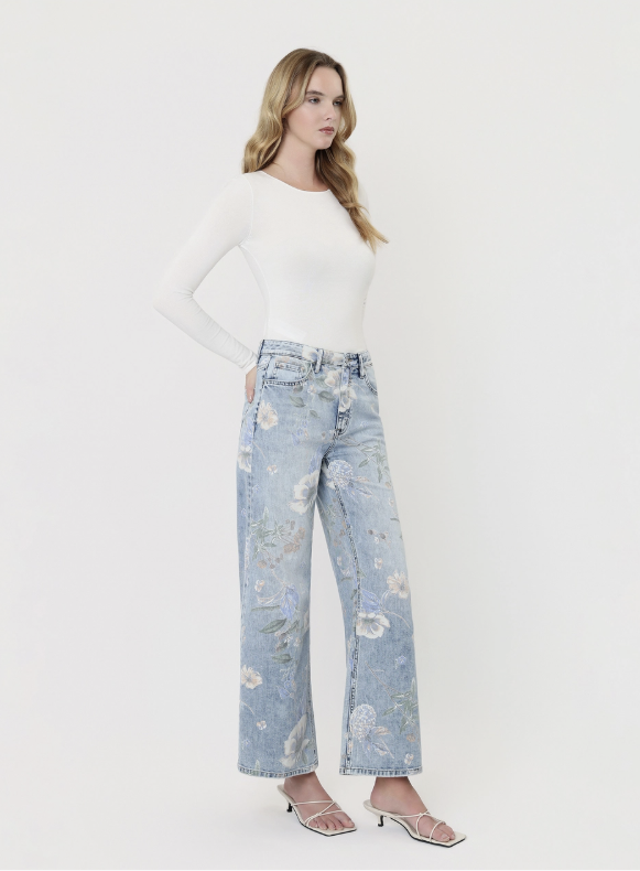 A woman modeling light-wash wide-leg jeans with a floral print, paired with a white long-sleeve top and strappy heels, against a plain off-white studio background.