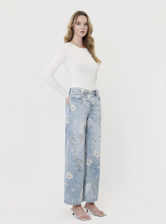 A woman modeling a white long-sleeve top and light blue wide-leg jeans with a floral print, paired with clear heeled sandals, against a plain white studio background.