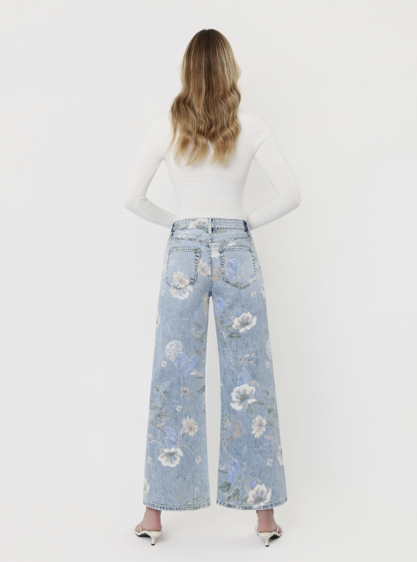 A woman viewed from behind wearing light blue wide-leg jeans with a white floral print, a white long-sleeve top, and white pointed-toe heels against a plain white studio background.