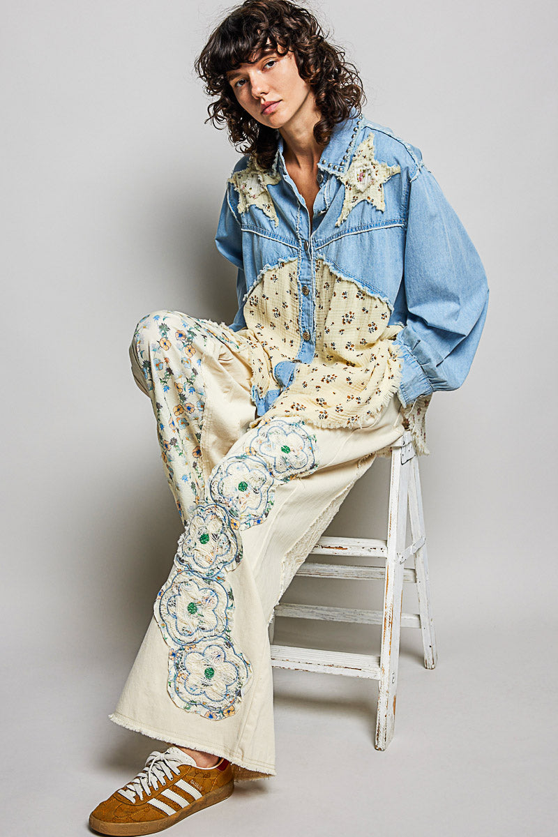 Person wearing a denim jacket and floral pants sitting on a stool against a plain background
