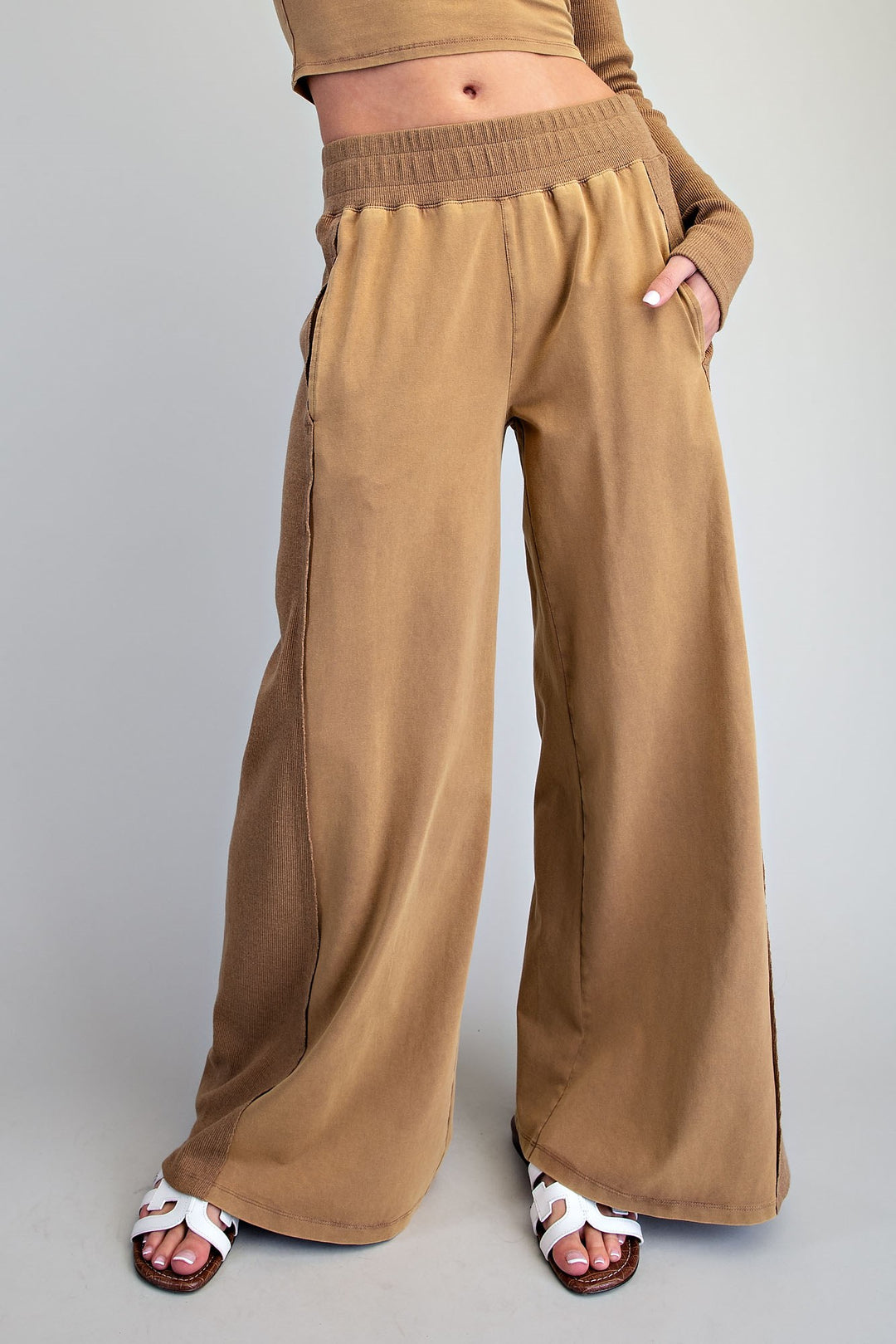 A person wearing wide-leg tan pants with an elastic waistband and side pockets, paired with white slide sandals, photographed against a plain light gray studio background.