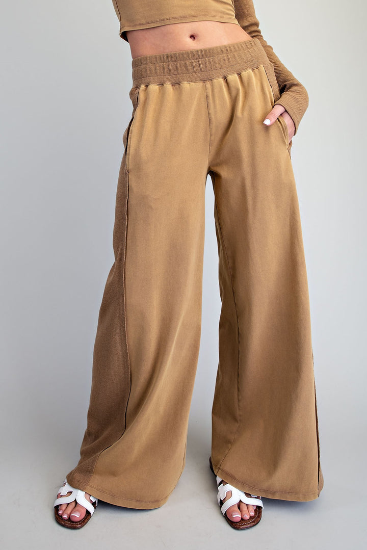 A person wearing wide-leg tan pants with an elastic waistband and side pockets, paired with white slide sandals, photographed against a plain light gray studio background.