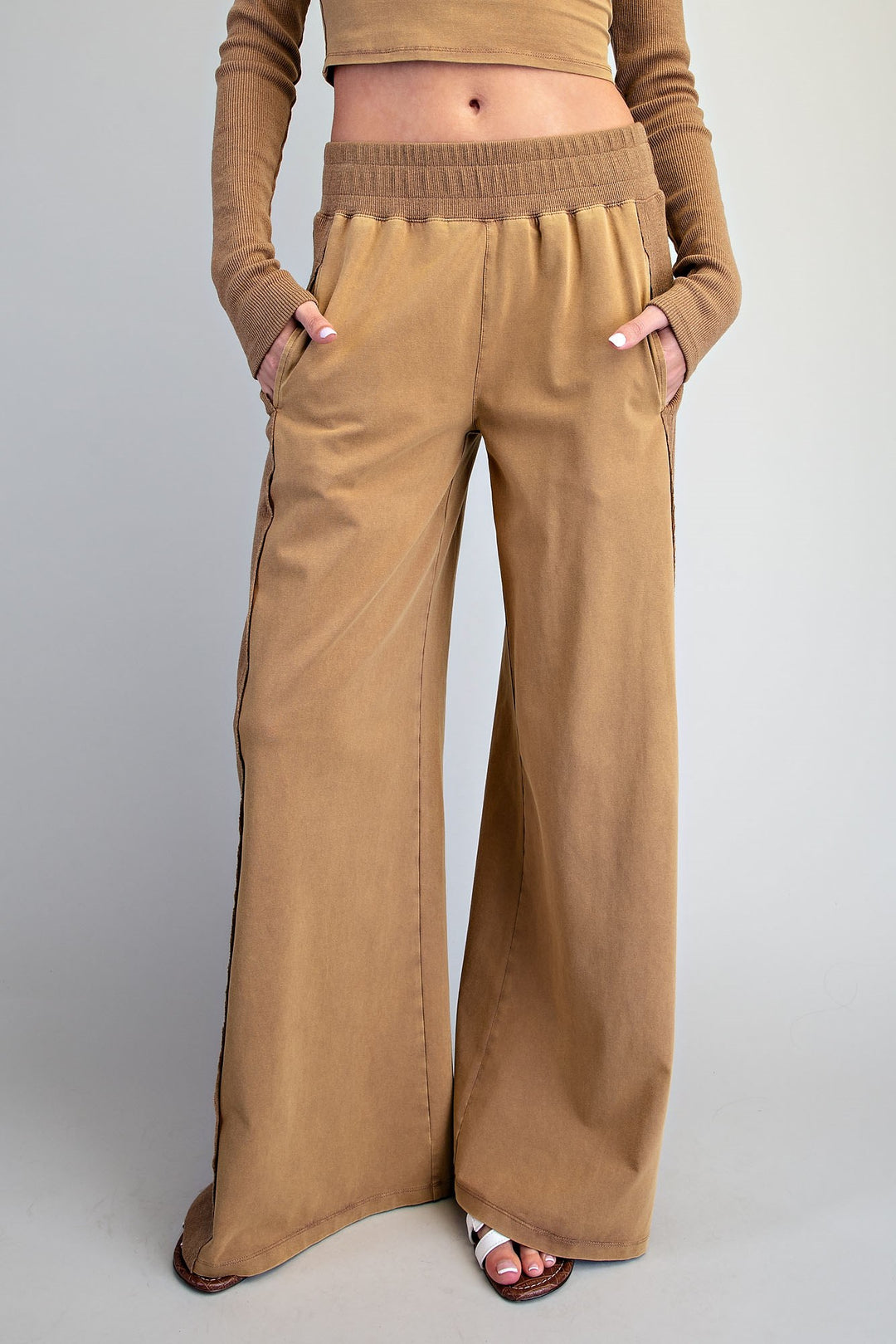 A model wearing wide-leg tan pants with side pockets and a matching ribbed waistband, paired with a matching tan long-sleeve crop top, against a light gray studio background.