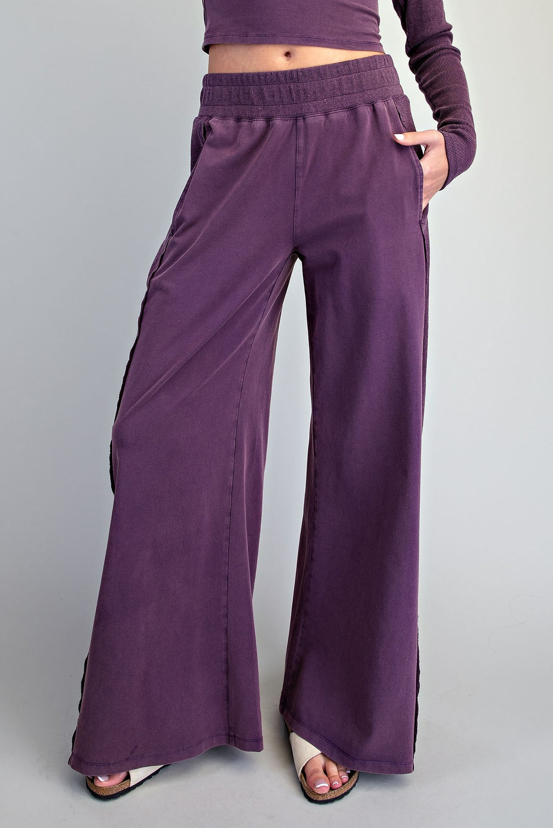 A model wearing a pair of wide-leg, deep purple lounge pants with side pockets and a matching purple long-sleeve crop top, shown from the waist down against a solid gray studio background.