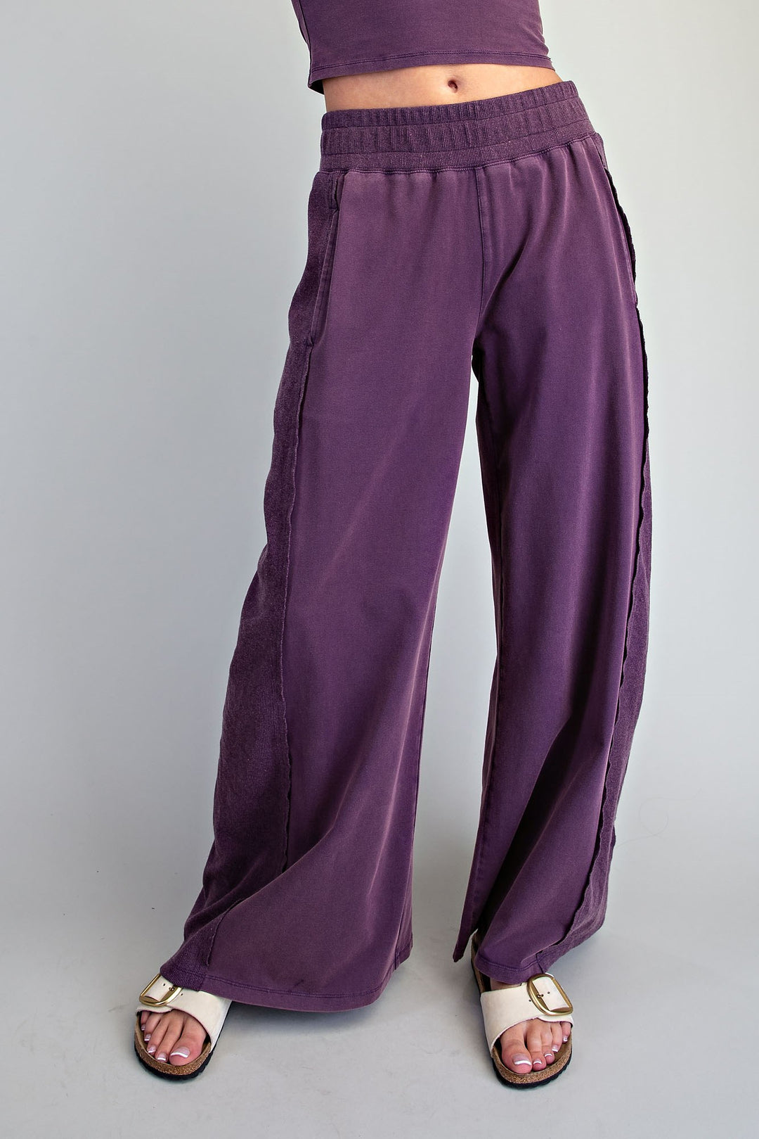 A person wearing loose, wide-leg purple pants with side seam detailing and white slide sandals, shown from the waist down against a plain light gray studio background.