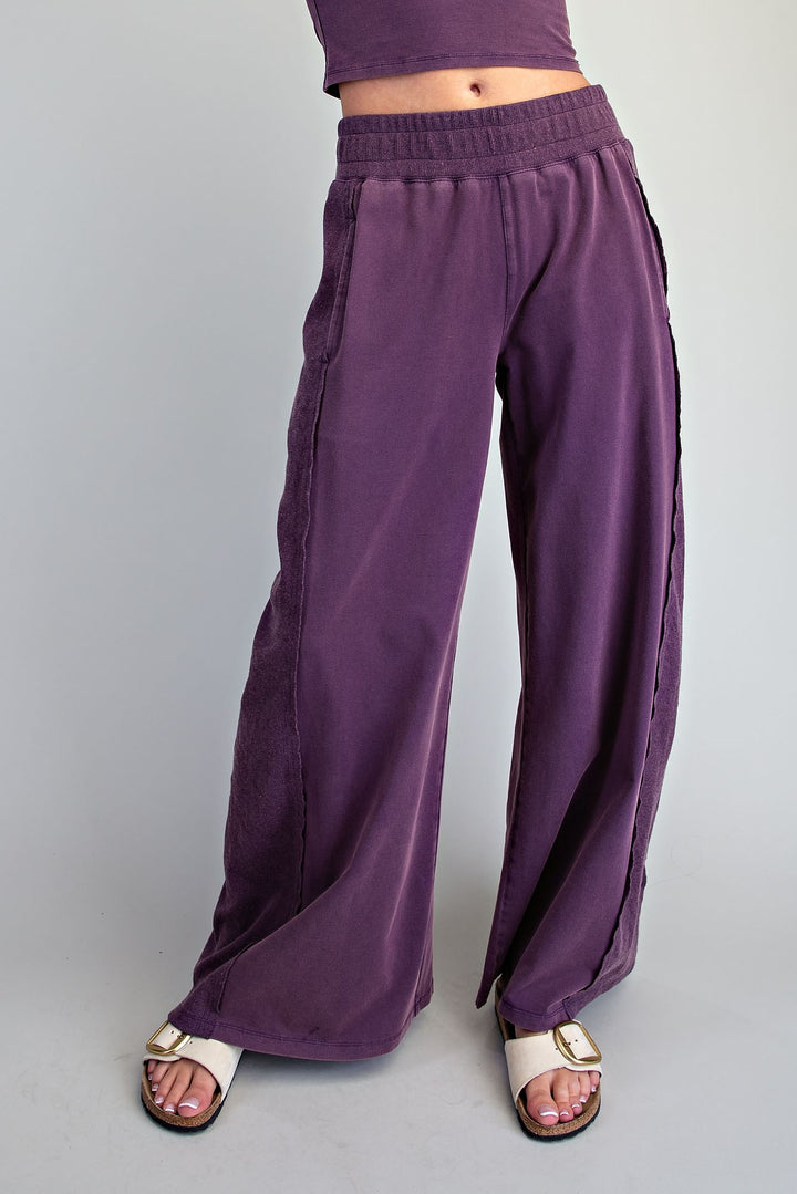 A person wearing loose, wide-leg purple pants with side seam detailing and white slide sandals, shown from the waist down against a plain light gray studio background.