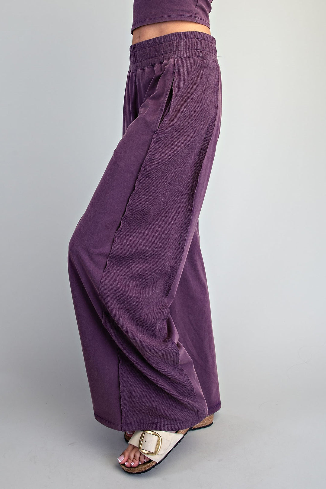 A person wearing wide-leg purple pants and white slide sandals, shown from the waist down against a light gray studio background.