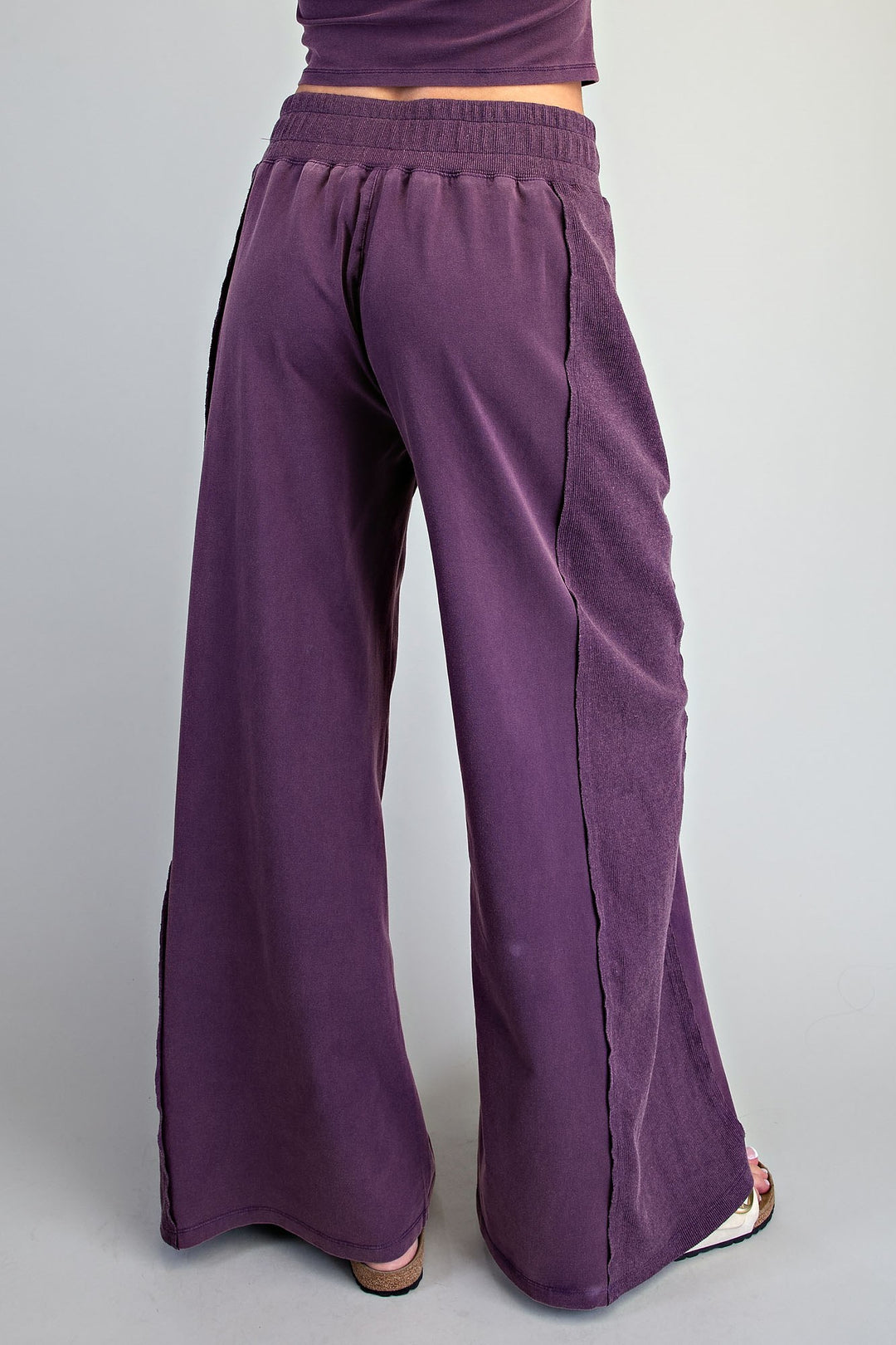 A person shown from the back wearing loose, wide-leg purple pants with an elastic waistband and side seam details, against a plain light gray studio background.
