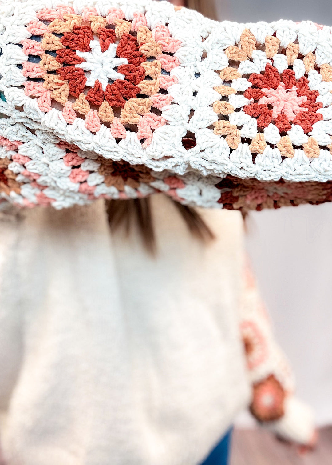 A close-up of a person wearing a chunky crocheted sweater with granny-square panels in cream, peach, rust, and white.