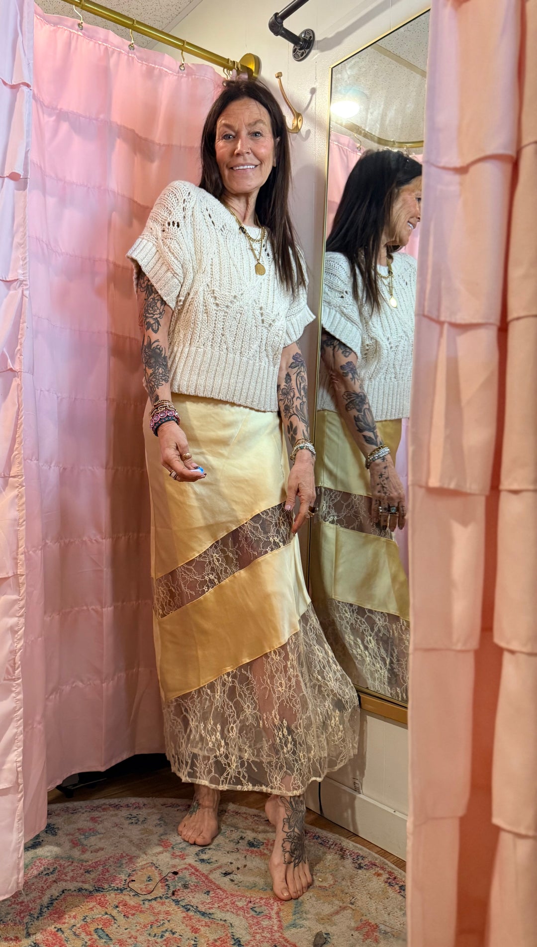 Person standing in a bathroom with pink shower curtain and patterned rug, wearing a white top and yellow skirt.
