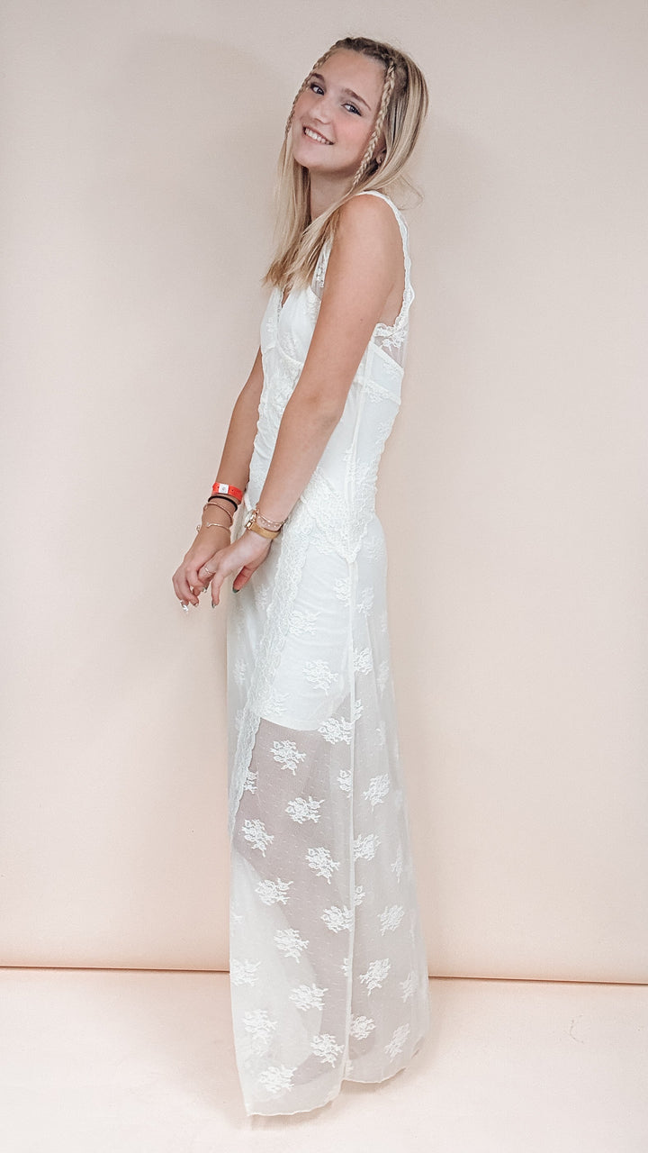 A woman modeling a sleeveless white lace dress with a sheer overlay skirt, standing against a solid pale peach background.