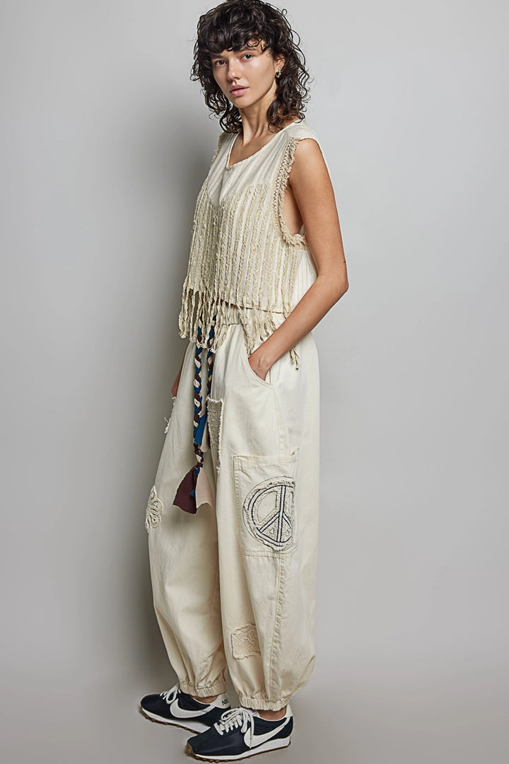 Woman wearing a beige sleeveless top and wide-leg pants with a peace symbol, standing against a plain background.