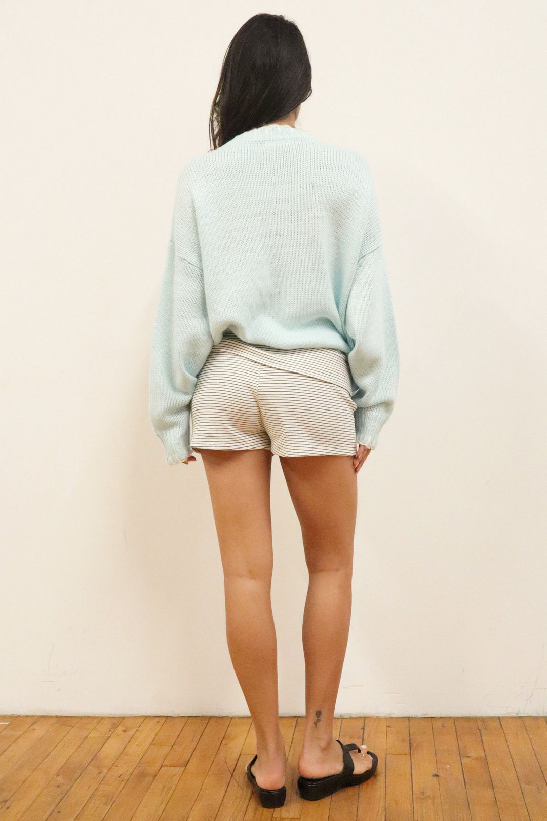 Person wearing a light blue sweater and white shorts standing against a plain wall.