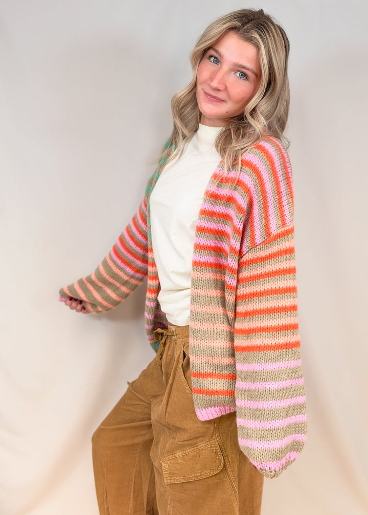 Woman shown from side view wearing colorful stripe cardigan with cream tee and camel color cord pants on plain background