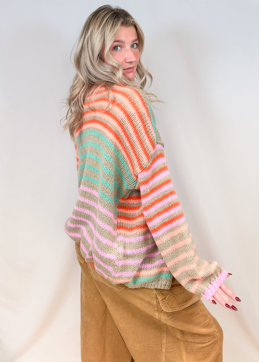 Woman shown from side view wearing colorful stripe cardigan with camel color cord pants on plain background