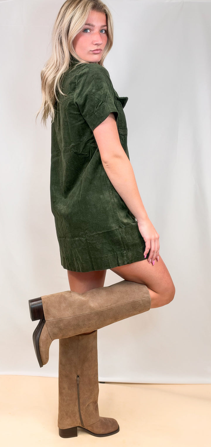 Woman shown from side view wearing green corduroy dress with knee high boots on plain background