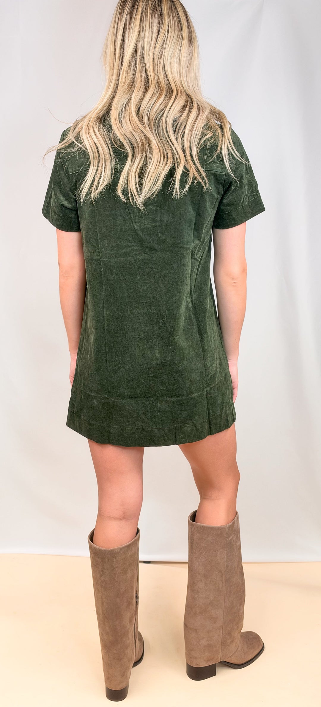 A woman shown from the back wearing a dark green corduroy short-sleeve dress and tall taupe-brown suede knee-high boots, standing against a white studio backdrop.