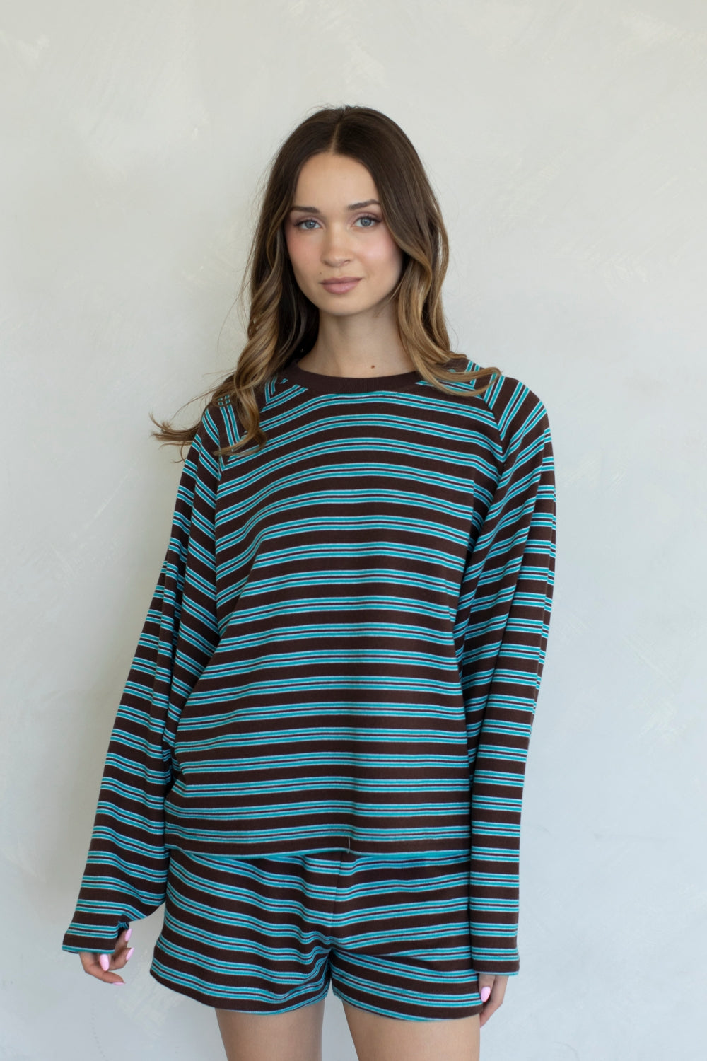 A woman modeling a long-sleeve striped top and matching shorts set. The set features horizontal stripes of teal, black, and thin brown lines, photographed against a light gray textured wall.