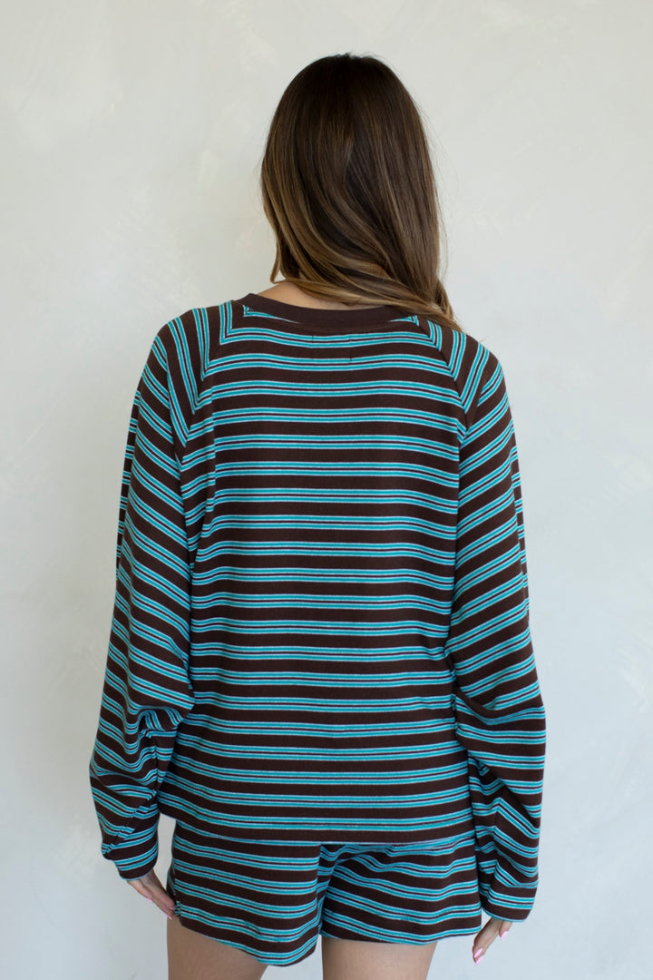 A woman viewed from behind wearing a loose long-sleeve top with horizontal teal, black, and brown stripes, paired with matching striped shorts, against a plain off-white wall.