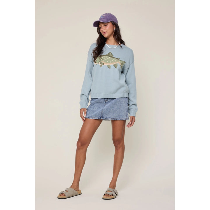 Woman wearing a light blue sweatshirt with a fish design, denim shorts, and a purple cap on a beige background.