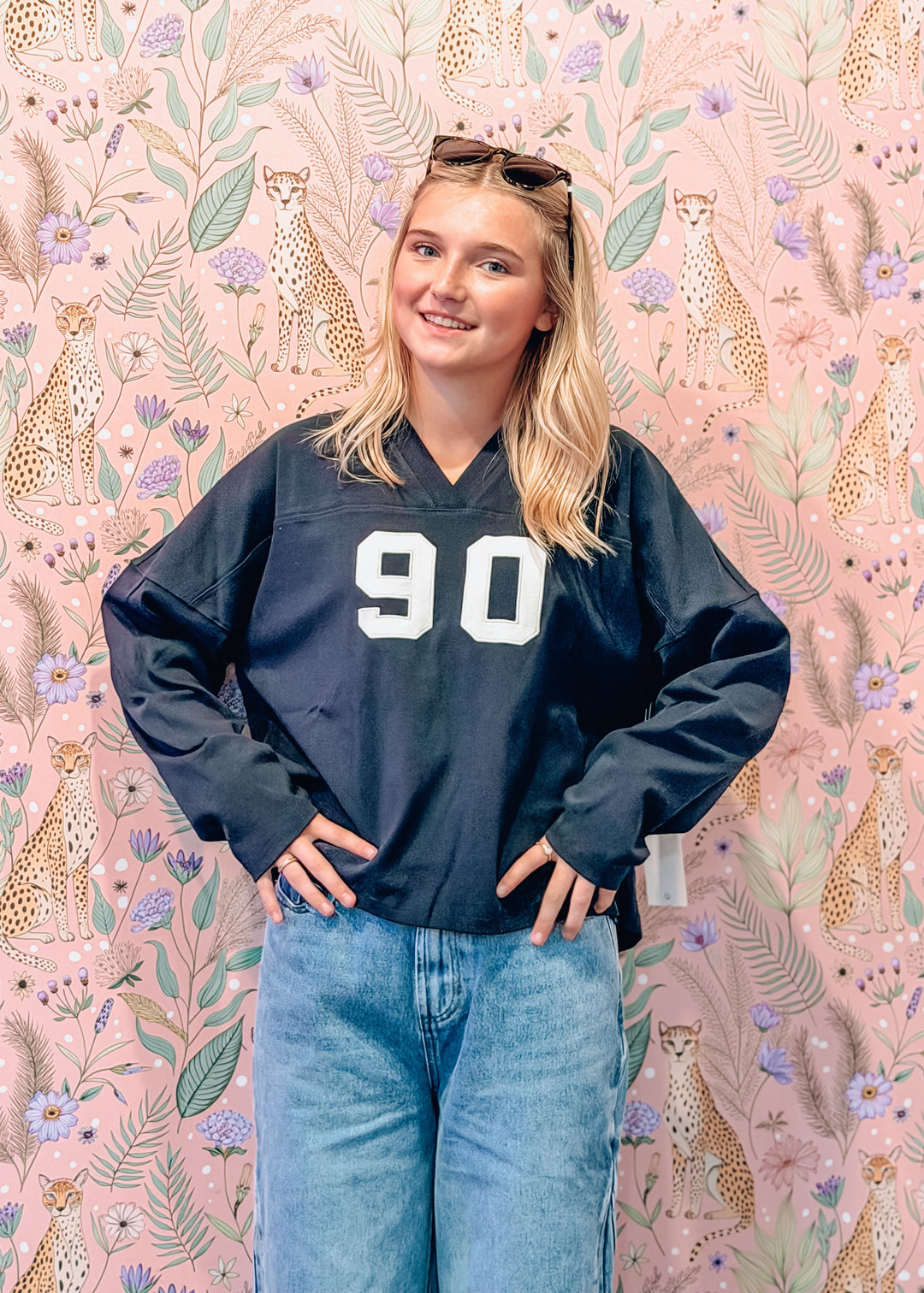 A young woman wearing a navy blue oversized v-neck jersey sweatshirt with the number 90 in white, paired with light blue jeans, standing in front of a pink floral and leopard print wallpaper.