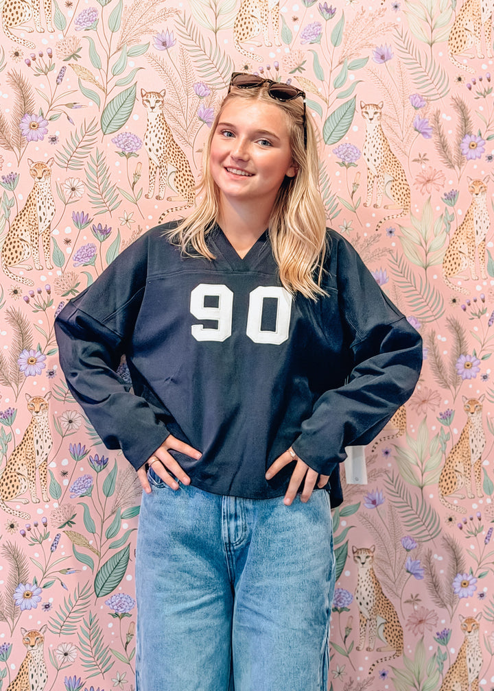 A young woman wearing a navy blue oversized v-neck jersey sweatshirt with the number 90 in white, paired with light blue jeans, standing in front of a pink floral and leopard print wallpaper.
