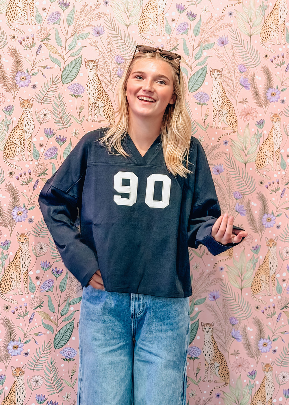 A smiling young woman stands in front of a pink floral and cheetah-patterned wallpaper, wearing a dark navy long-sleeve V-neck jersey with the number 90 in white on the front, paired with light blue jeans.