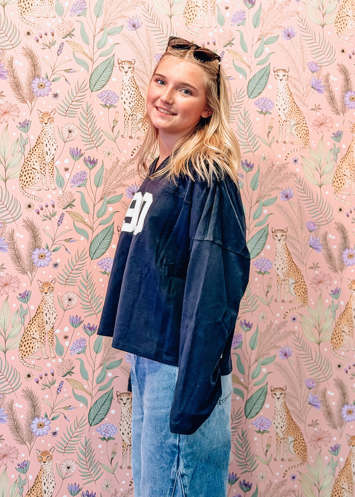 A young woman with blonde hair and sunglasses on her head stands in front of a pink floral and cheetah-patterned wallpaper. She is wearing a dark navy blue oversized top with a white number &